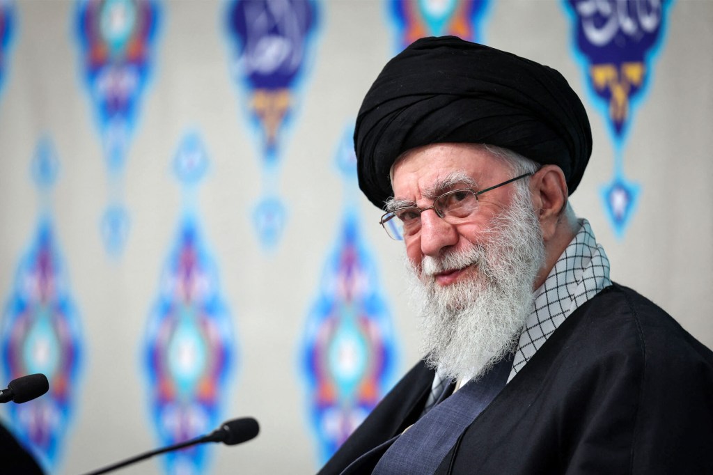 Ayatollah Ali Khamenei sitting in front of a patterned background with two microphones in front of him.