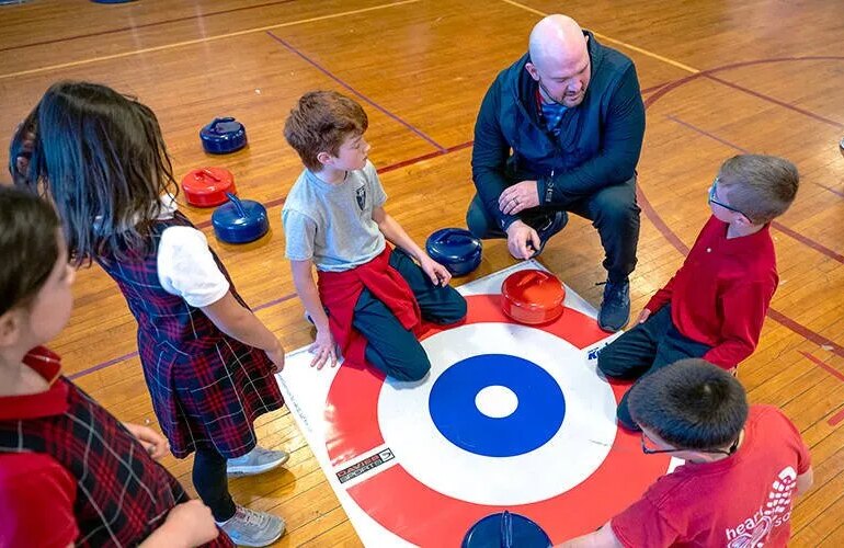 A man kneels down to talk to students in a gym.