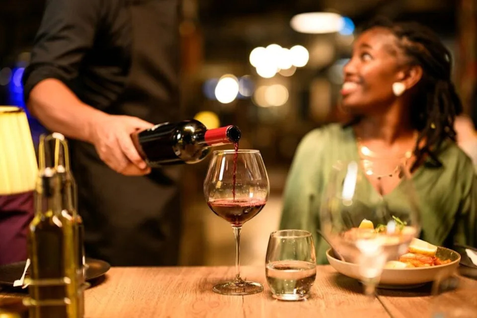 sommelier pouring wine at dinner table with Black woman as dining guest