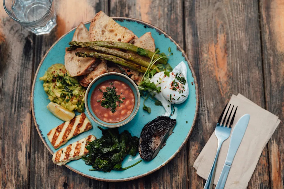 A plate of full vegetarian breakfast with meshed avocado, halloumi cheese, poach egg, grilled asparagus, bread and mushroom served on a wooden dining table at restaurant.