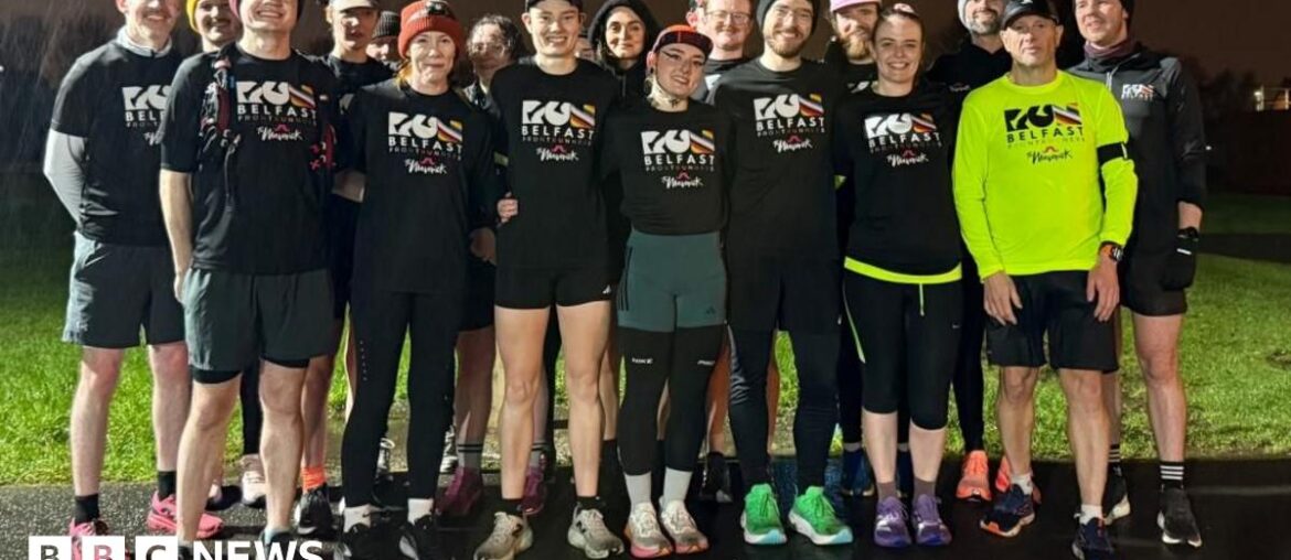 A group of runners standing together. It is raining and night time. Most of them are wearing black T-shirts which say "Run Belfast" on them in white lettering, one man to the front left of the group has the same logo but in black text on a neon yellow running top.