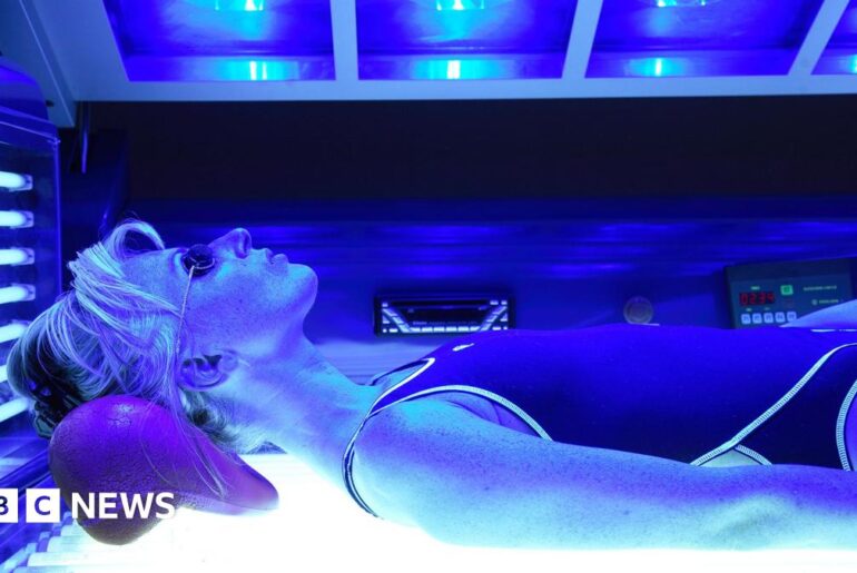 A woman lies on her back in a sunbed, wearing goggles - tubes surround her emitting blue light.