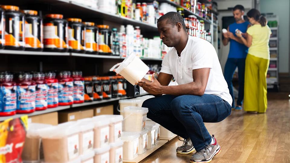 A man crouches next to shop shelves filled with sports supplements, while looking at one container of cream-coloured powder.