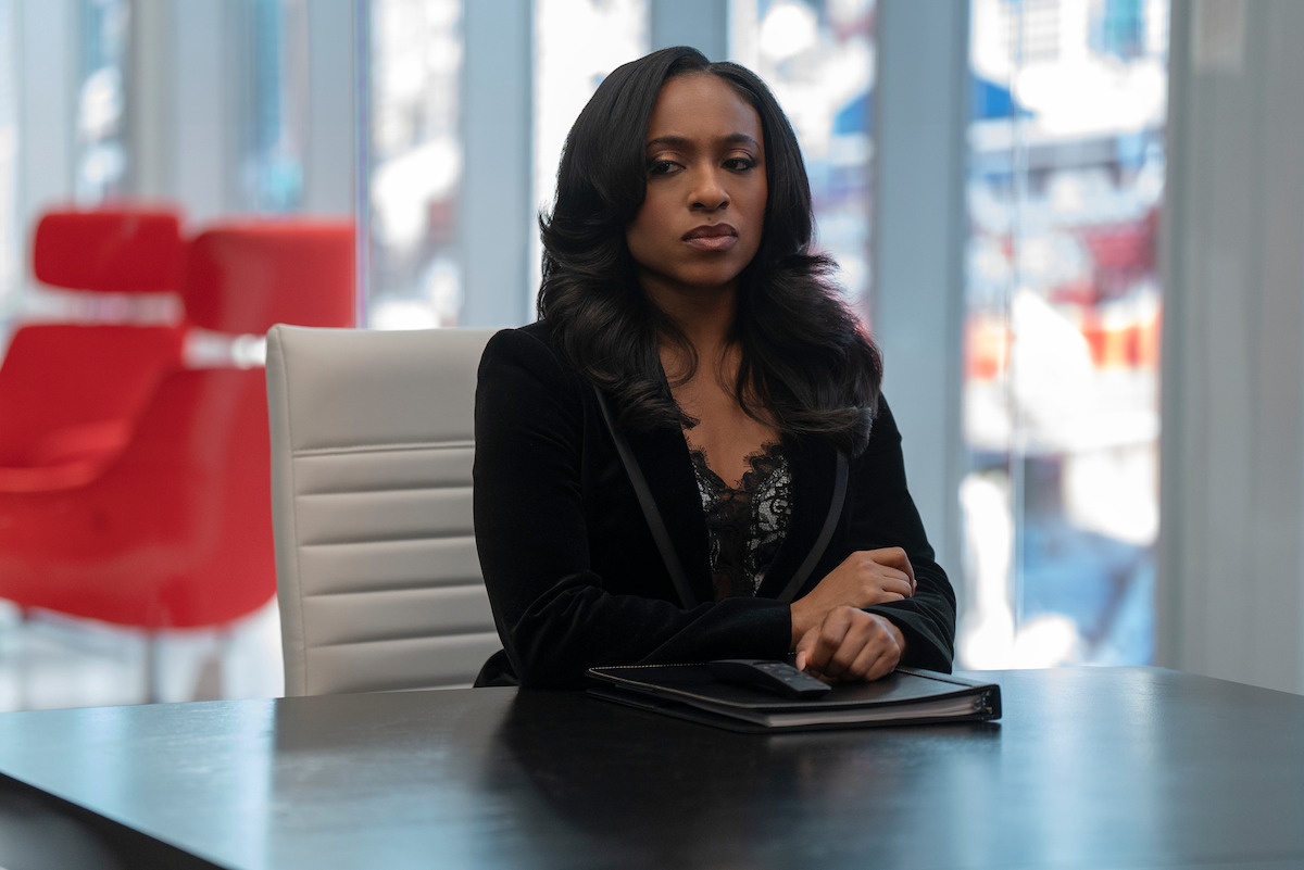 Taylor Polidore Williams as Kimmie in black blazer sitting at a modern office table, holding a folder, with large windows and red chairs in the background, bright daylight streaming in.