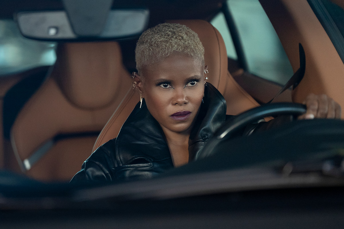 Jasmine Burke as Gina with short blond hair and dark lipstick, wearing a black leather jacket, sits in the driver's seat of a car with tan leather interior, gripping the steering wheel and looking ahead with a focused, intense expression.