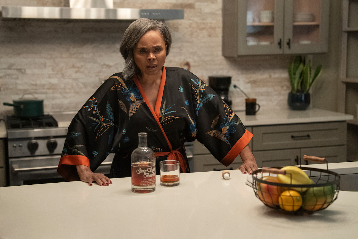Debbi Morgan as Olivia wearing a black floral robe stands in a modern kitchen, leaning on a counter with a bottle and glass of liquor; there is a fruit basket and stove nearby, and the mood feels tense.
