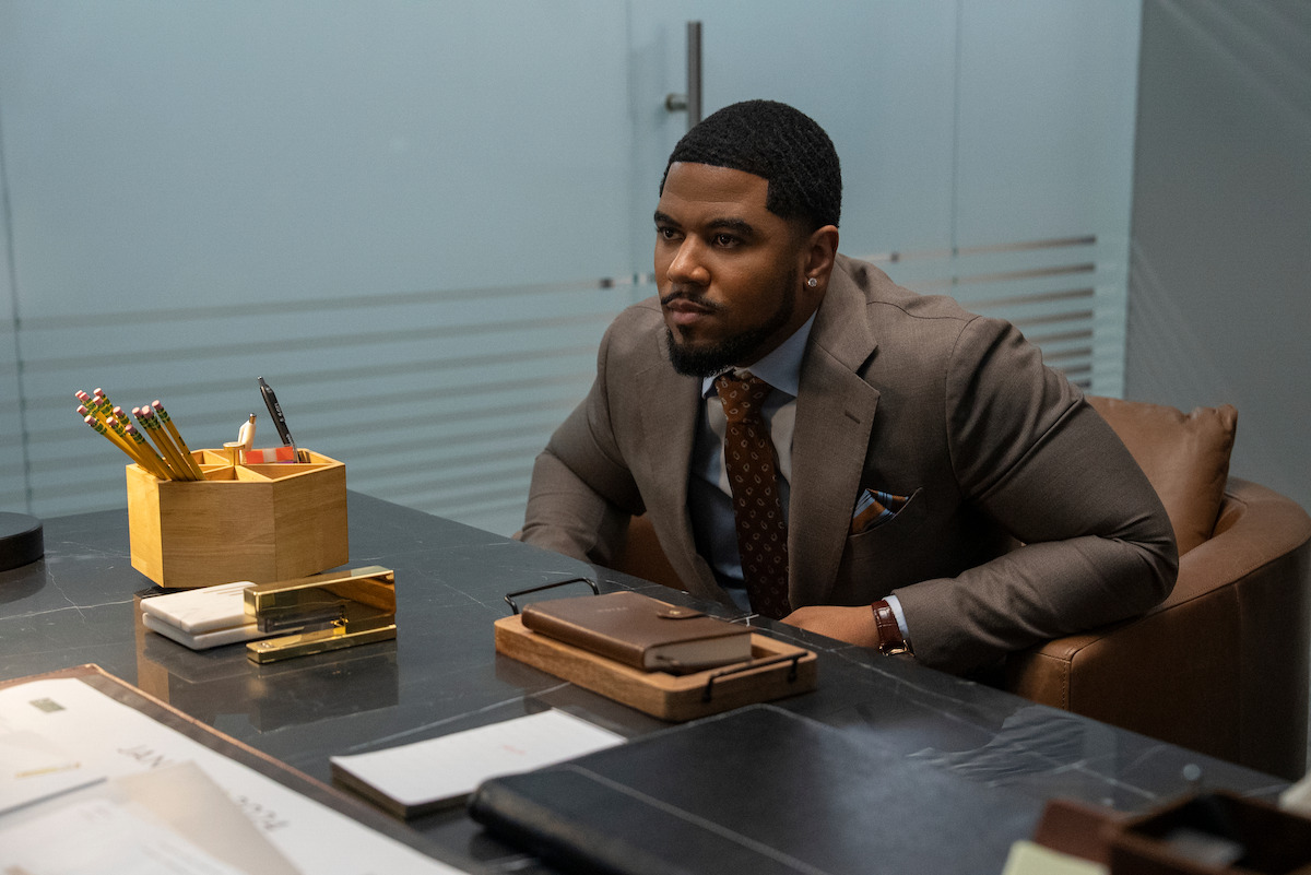 Julian Horton as Roy in a suit sits at a modern office desk with organized stationery, notebooks, and papers; glass walls and a door suggest a professional, corporate environment.