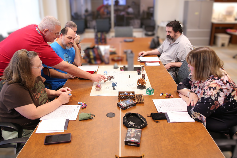 Six people engage in a tabletop role-playing game, Dungeons & Dragons, gathered around a large wooden table with maps, dice, and miniatures.