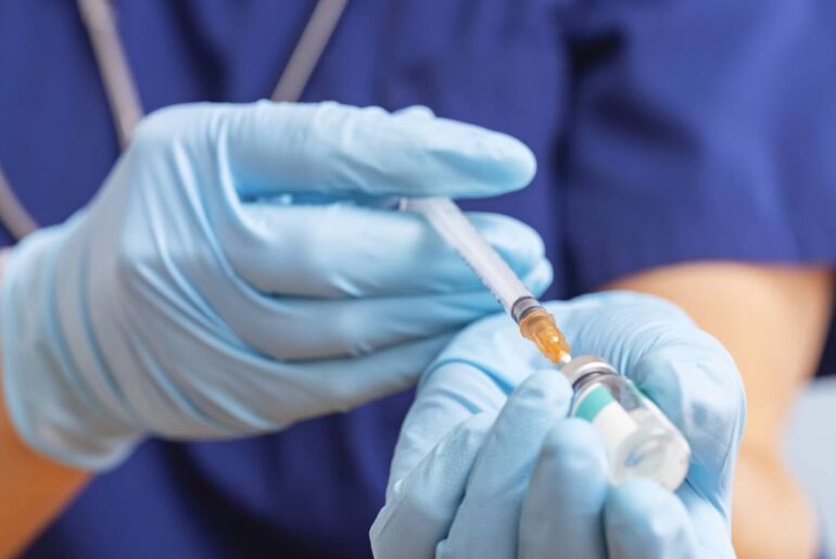 A pair of gloved hands fills a syringe from a vial of vaccine.