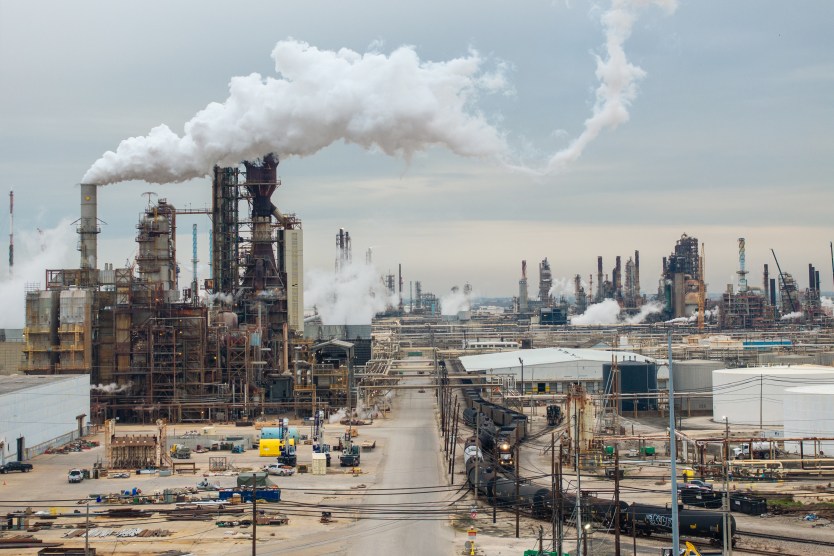 Road cuts through large refinery on a flat landscape. Smoke billows from tall machinery against a grey sky.