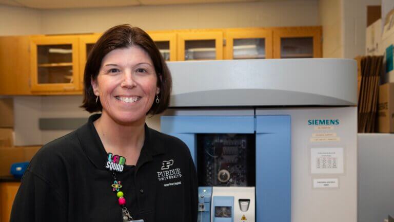 A woman standing in front of a piece of laboratory equipment, she is wearing a black shirt and a colorful lanyard
