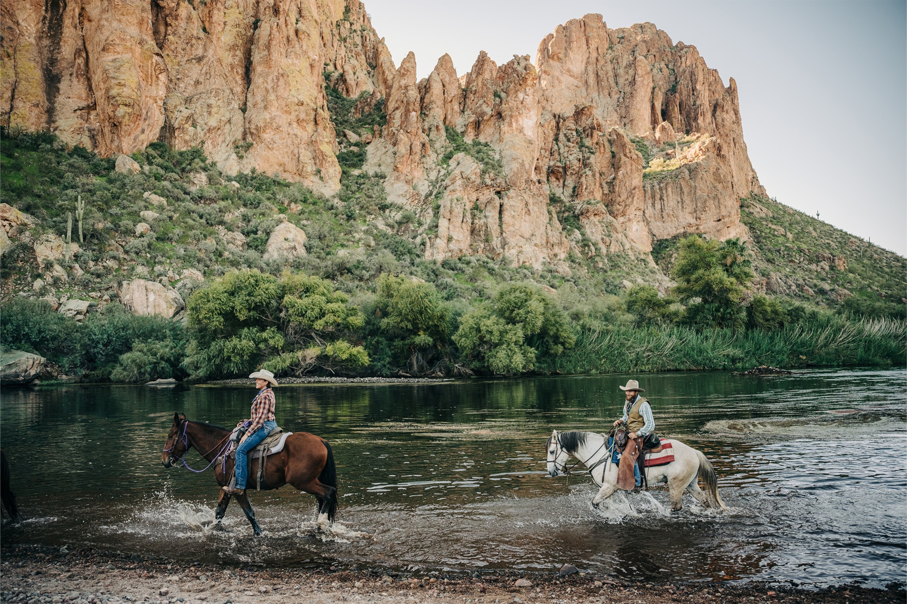 Two wranglers on horseback ride through the shallow river