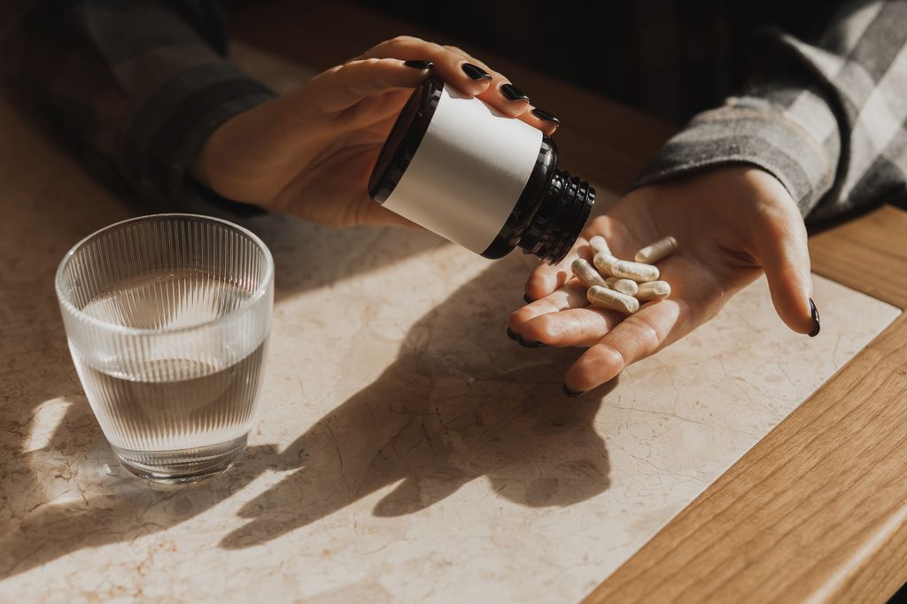 A close-up scene shows a person pouring white vitamin capsules from a bottle into an open palm beside a glass of water, highlighting health, supplements, and daily wellness routines.