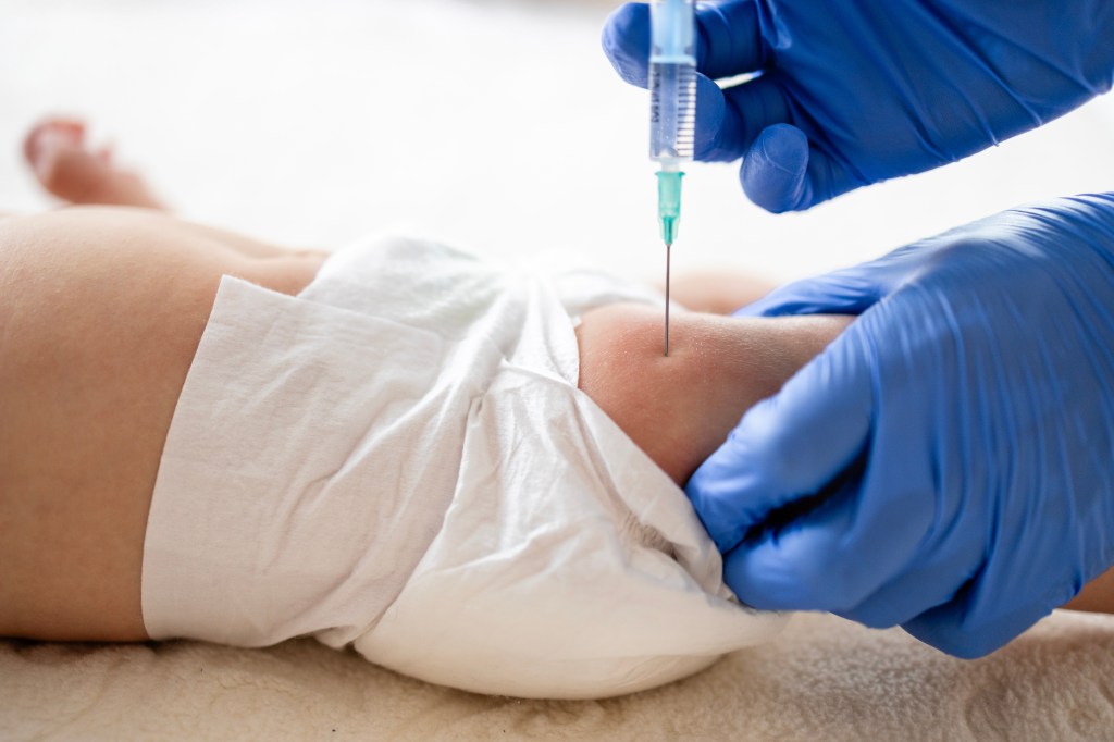 A healthcare worker in blue gloves injects a vaccine into a baby's thigh.