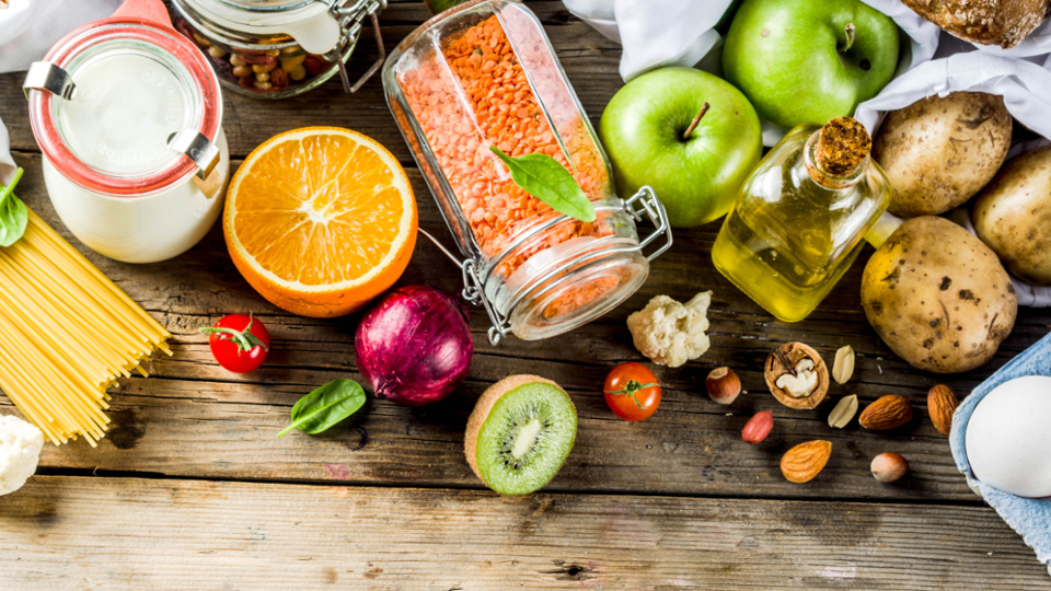 Ansicht von oben auf einen Tisch mit Obst, Gemüse, Öl, Nudeln, Nüsse und ein Ei