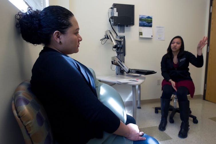 A pregnant woman sits in a clinic exam room while a health care provider talks to her.