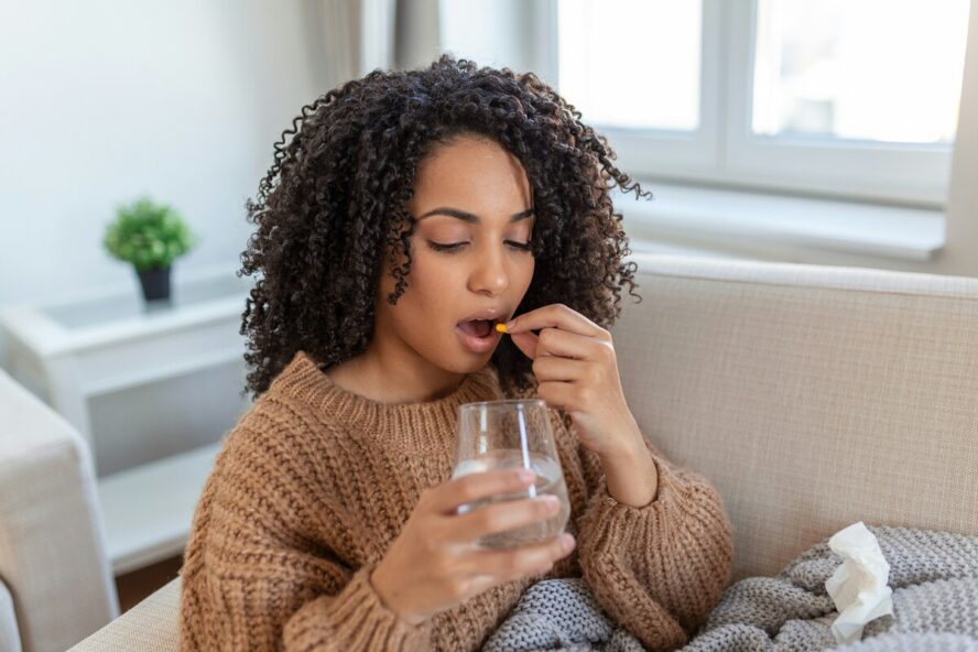 Image of a young woman sitting on a couch and taking a supplement with a glass of water in her hand.