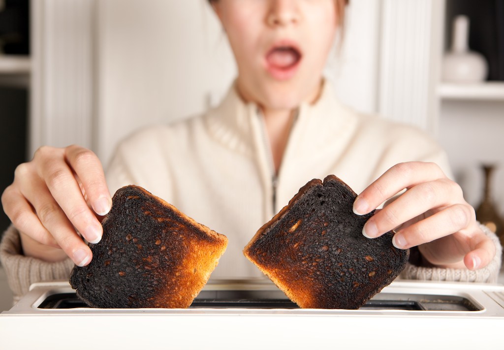 A woman with her mouth open in surprise holds two slices of burnt toast from a toaster.