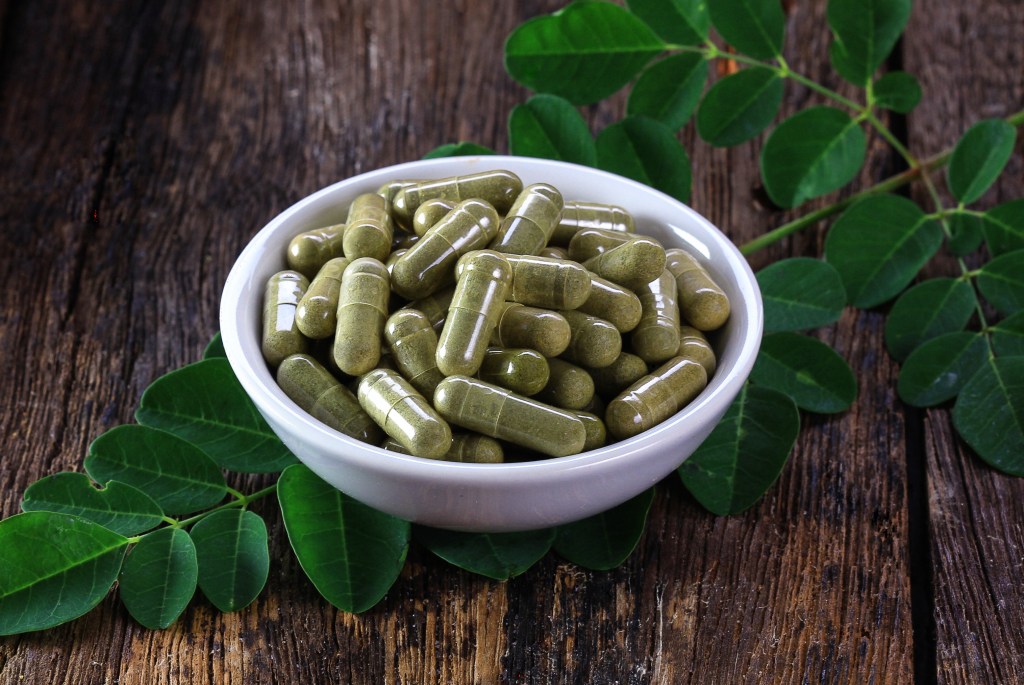Herbal capsules made from moringa leaf in a white bowl on a wooden surface with green leaves.