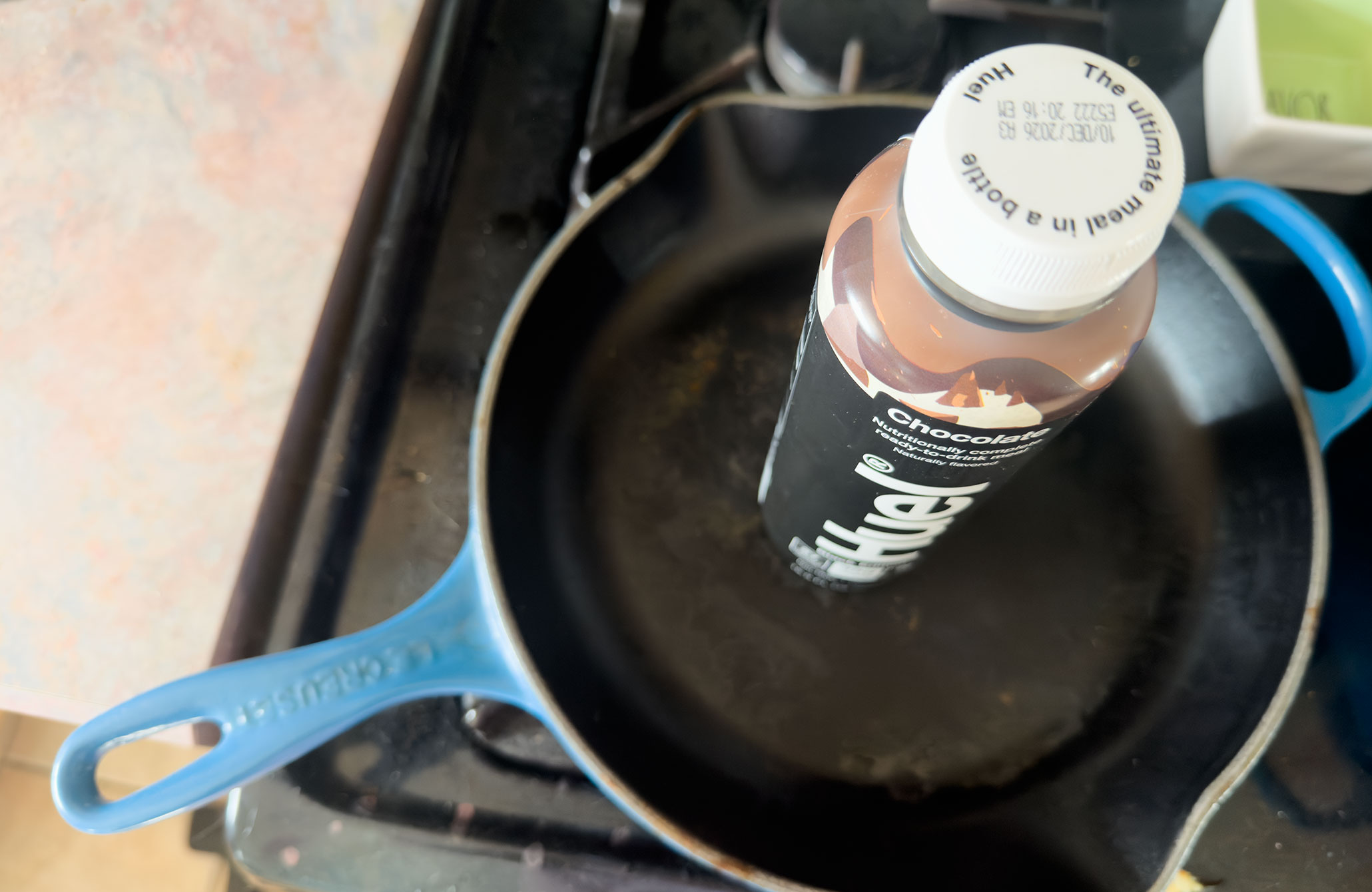 A Huel bottle where the top of the cap reads ‘The ultimate meal in a bottle.’ The bottle sits on top of a frying pan above a stove.