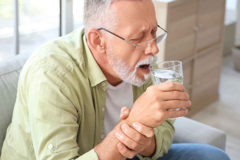 Older man drinking water while coughing, illustrating Parkinson’s related swallowing and throat symptoms.