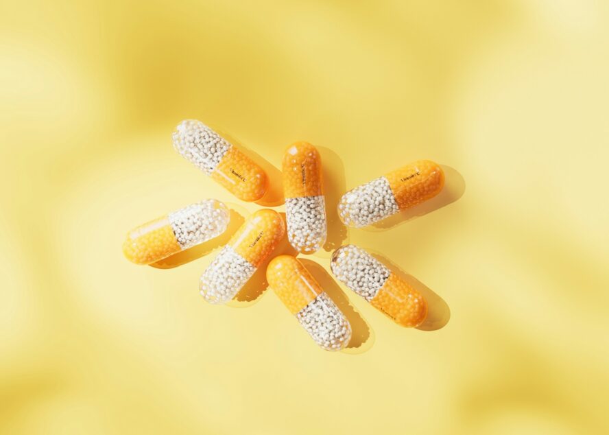 Image of seven clear capsules with a split of tiny orange and white beads inside, set against a yellow background.