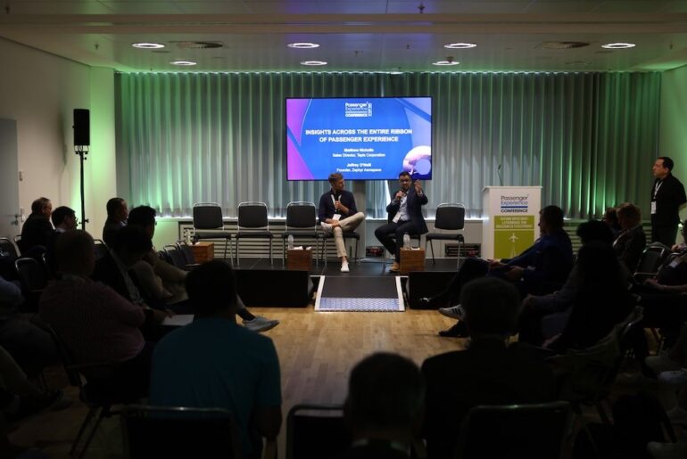 Conference panel discussion with two speakers seated on stage in front of a projected presentation slide titled ‘Insights Across the Entire Ribbon of Passenger Experience’ with green ambient lighting and seated audience