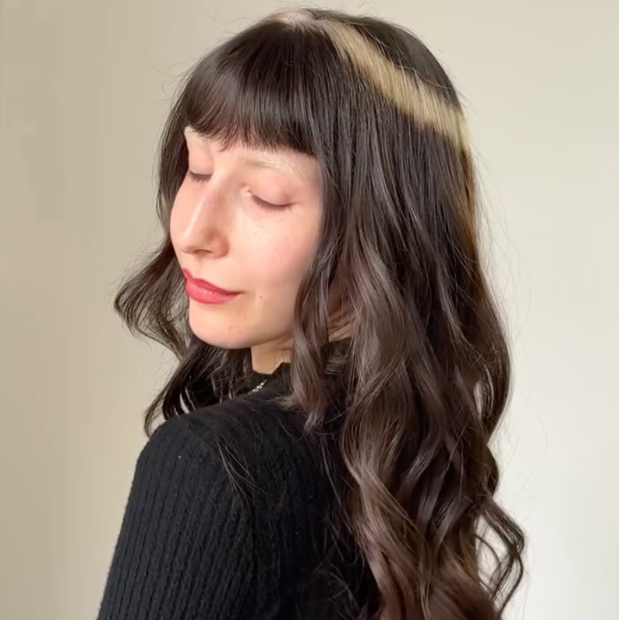 person with long wavy hair wearing a black ribbed top and belt standing sideways