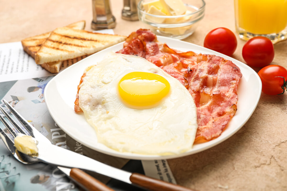Photo shows a white plate loaded with bacon and eggs, with butter and a few tomatoes on the side