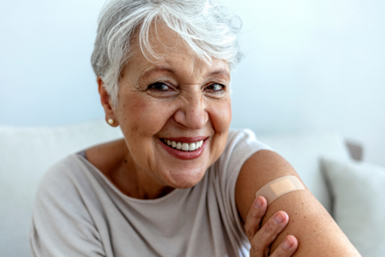 A smiling woman with short white hair shows off a bandaid on her upper arm.