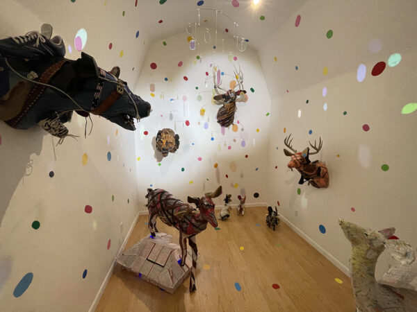 A photograph of an installation by Ken Little and Cathy Cunningham-Little featuring sculptured animal heads hung on the walls and animal forms placed on the floor with small colorful mirrored circles throughout the space.