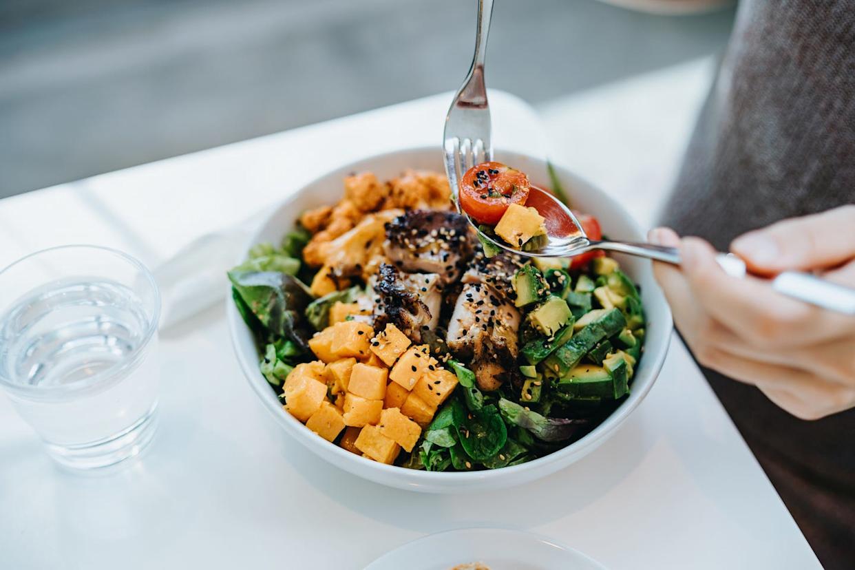 foods linked to longevity close up, high angle shot of young woman enjoying multi coloured healthy fruit, vegetables with grilled chicken salad bowl with balanced nutrition in cafe, with a glass of water by the side. healthy eating lifestyle. people, food and lifestyle concept