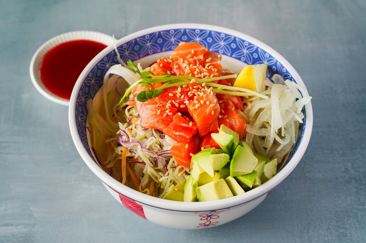 A bowl of teriyaki salmon topped with fresh salad, avocado, and onion, accompanied by a savory dipping sauce.