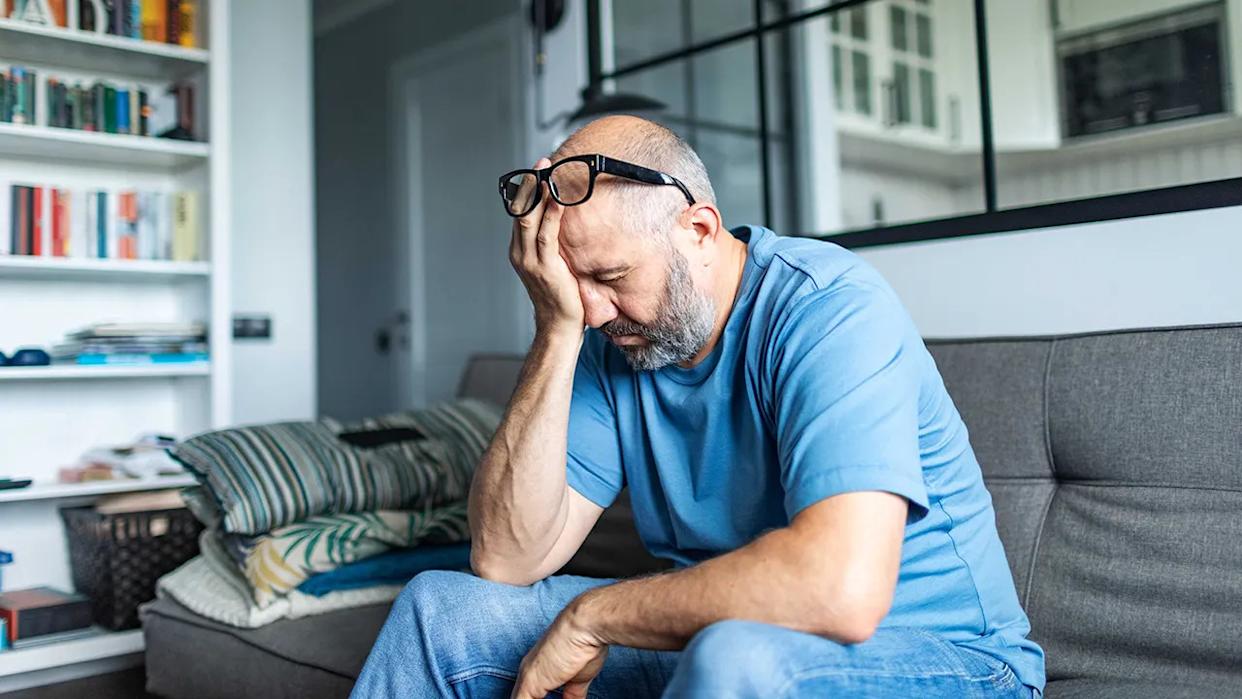 Middle-aged man sitting on a couch with his head in his hand, looking stressed or fatigued at home.