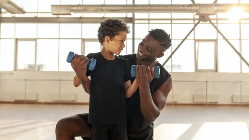 African American boy working out and lifting dumbbells in the gym with his dad, man training his son and helping him