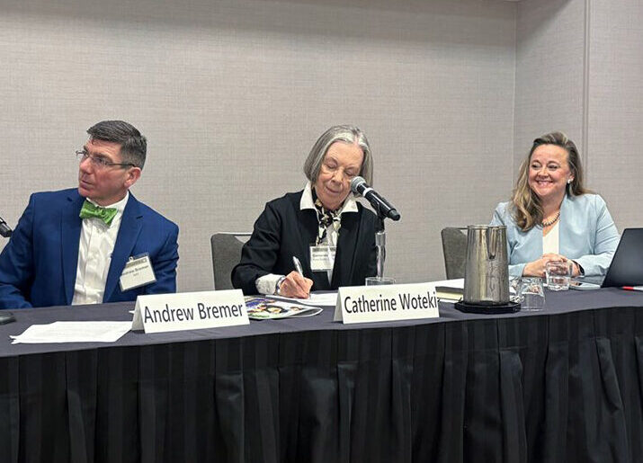 Four people in professional attire seated at a conference table with microphones and name tags on the table for each panelist