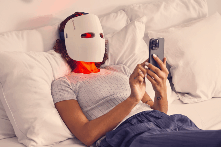 A person wearing a white LED light therapy mask with red lights glowing, lying on a bed with white pillows and sheets, while looking at a smartphone.