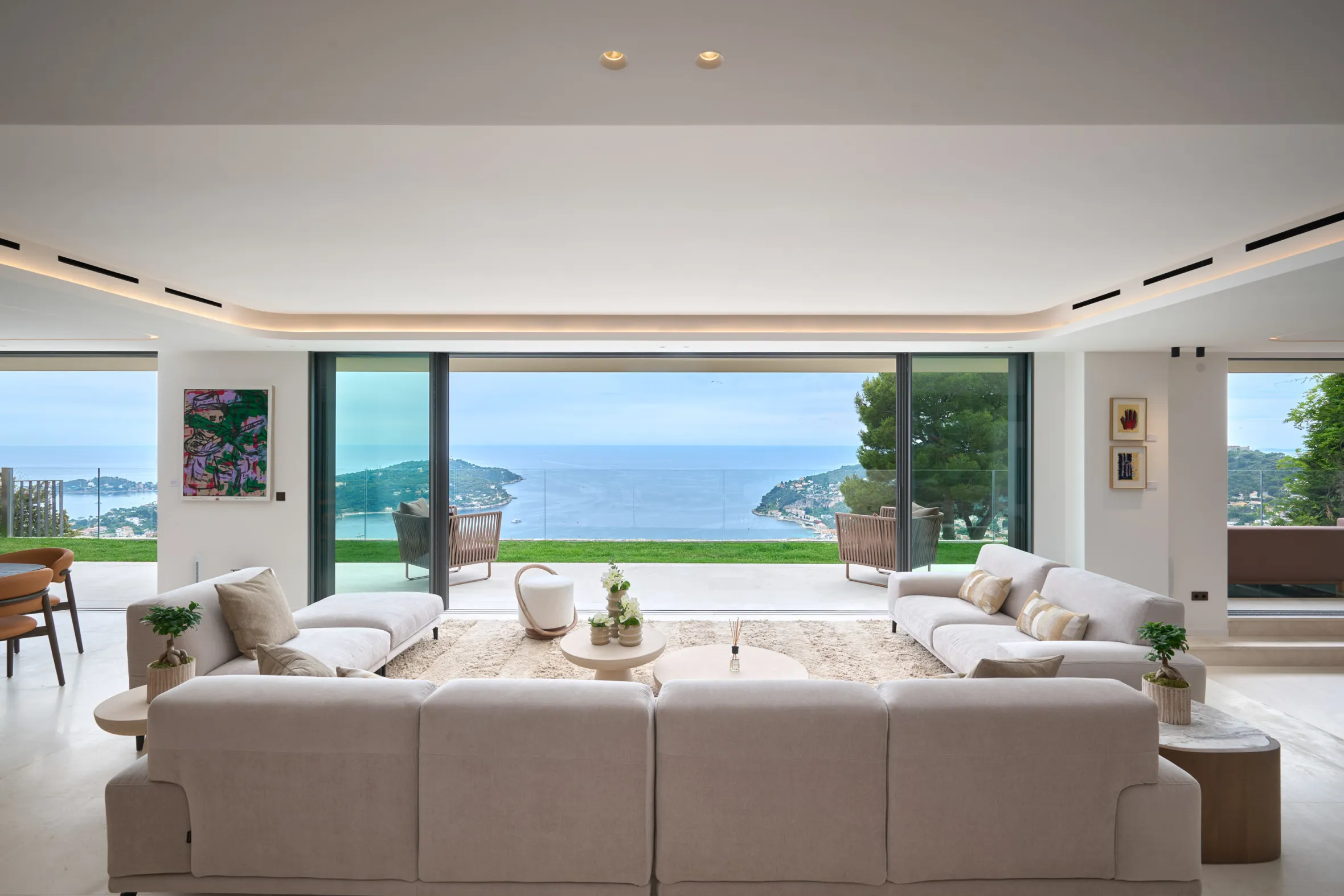 A luxurious living room with light-colored sofas facing a large glass wall that opens onto a patio overlooking the sea.