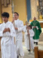Boys in white robes process in a church, holding a cross and candles. A priest in a green robe follows, with an ornate altar in the background.