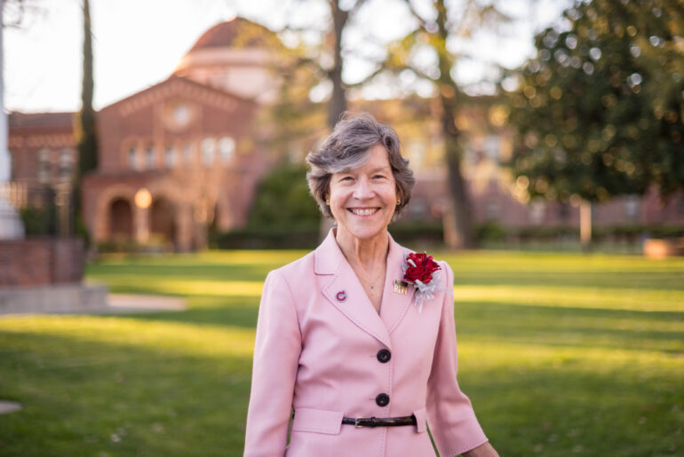 A woman wearing a light pink blazer with a red boutonniere stands smiling on a grassy campus lawn. Trees and a brick building with arched windows and a domed roof appear softly blurred in the background in warm afternoon light.
