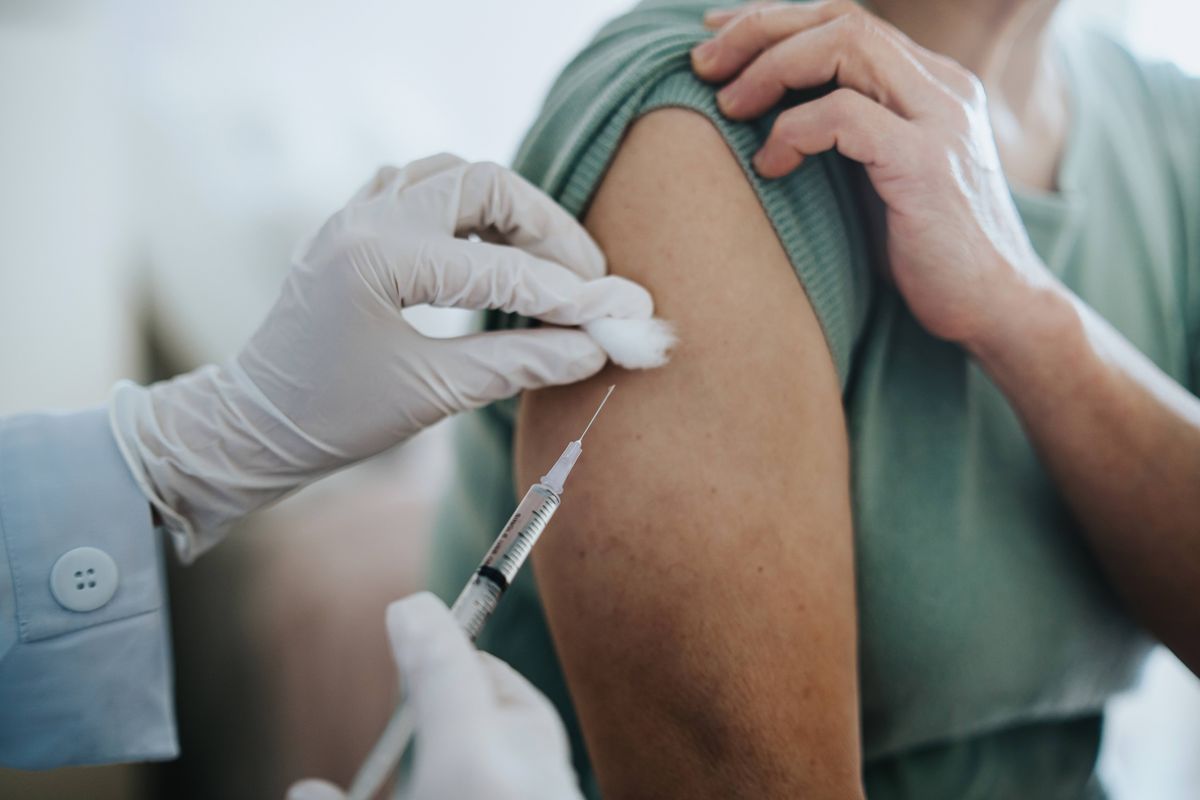 A healthcare professional administers a vaccine to a patient's upper arm. The individual administering the vaccine is wearing gloves and a medical gown, while the patient is dressed in a green top. The scene takes place in a clinical setting.