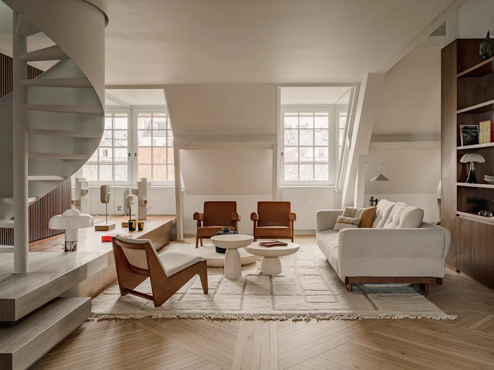 A refined attic apartment interior characterized by a warm, modernist aesthetic. The space is organized as an open-plan living area under a pitched roof, with exposed white-painted beams and triangular dormer windows that bring in soft natural light.