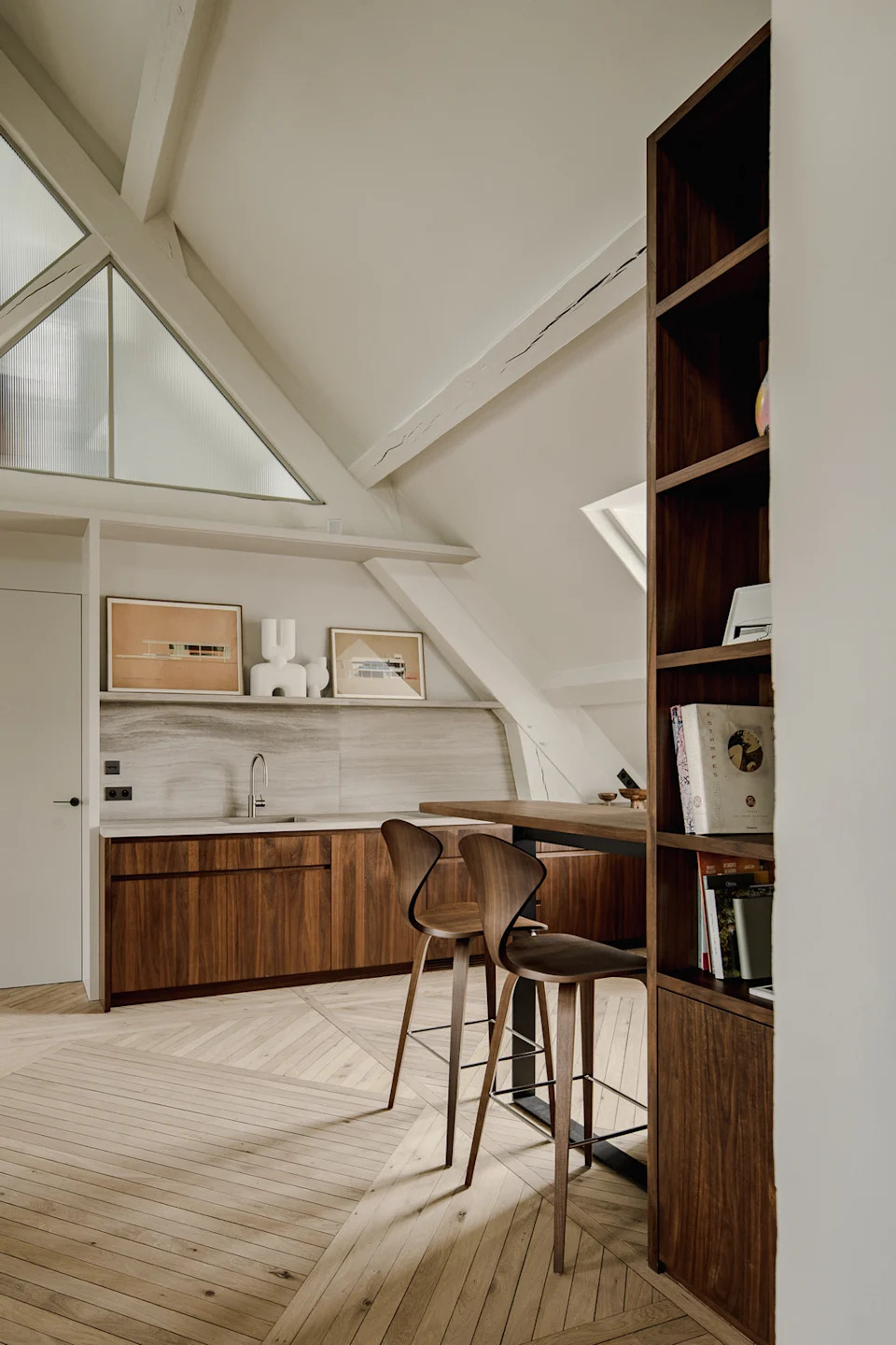 A refined attic apartment interior characterized by a warm, modernist aesthetic. The space is organized as an open-plan living area under a pitched roof, with exposed white-painted beams and triangular dormer windows that bring in soft natural light.