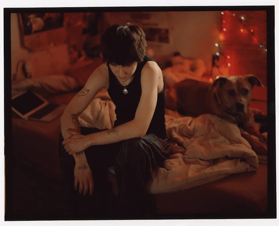 Kaleb sits on a bed beside a dog in a dimly lit room.