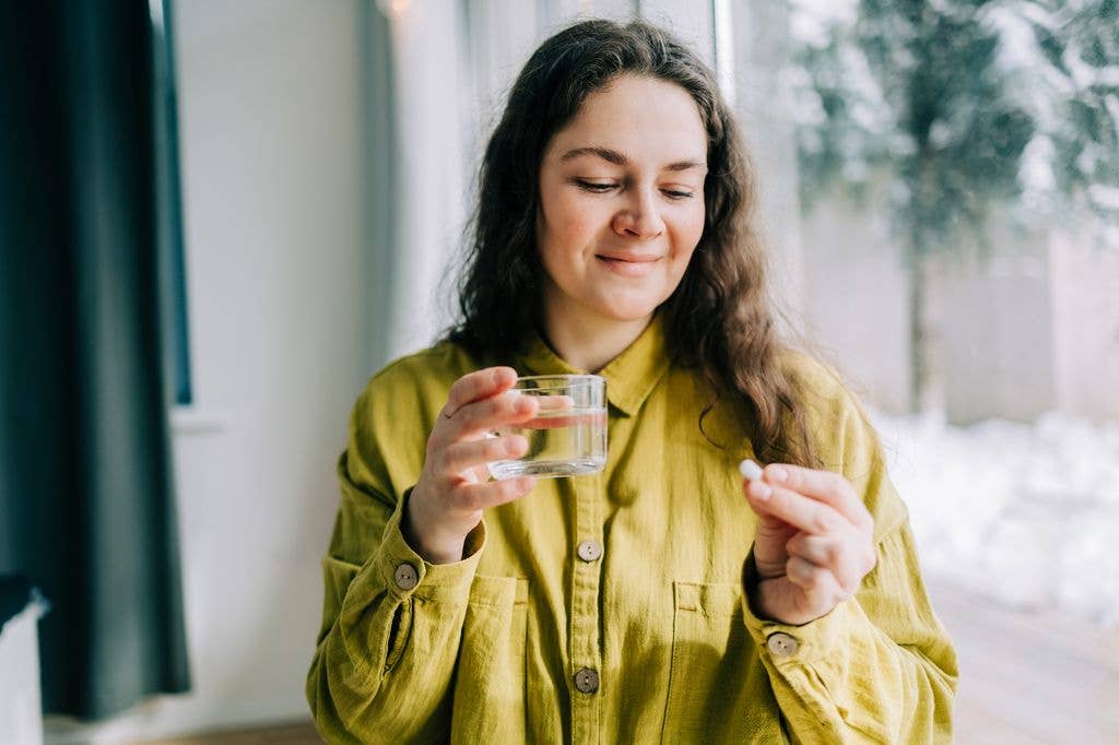 Smiling woman holding pill and glass of water, taking medicine at home