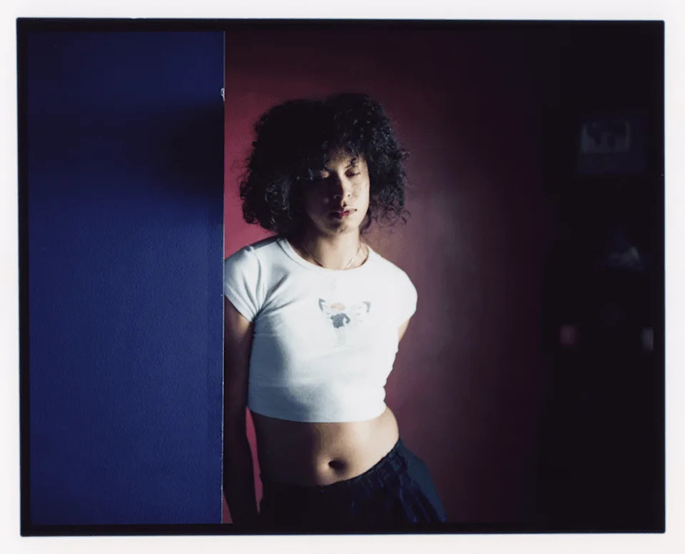 Quartz stands against a wall in a dimly lit room, wearing a white cropped T-shirt