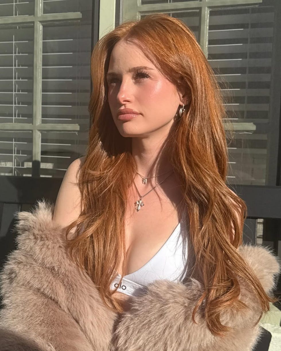 Person with long red hair wearing a white top and a fur shawl, sitting in a sunlit environment. Person with long red hair wearing a white top and a fur shawl, sitting in a sunlit environment.