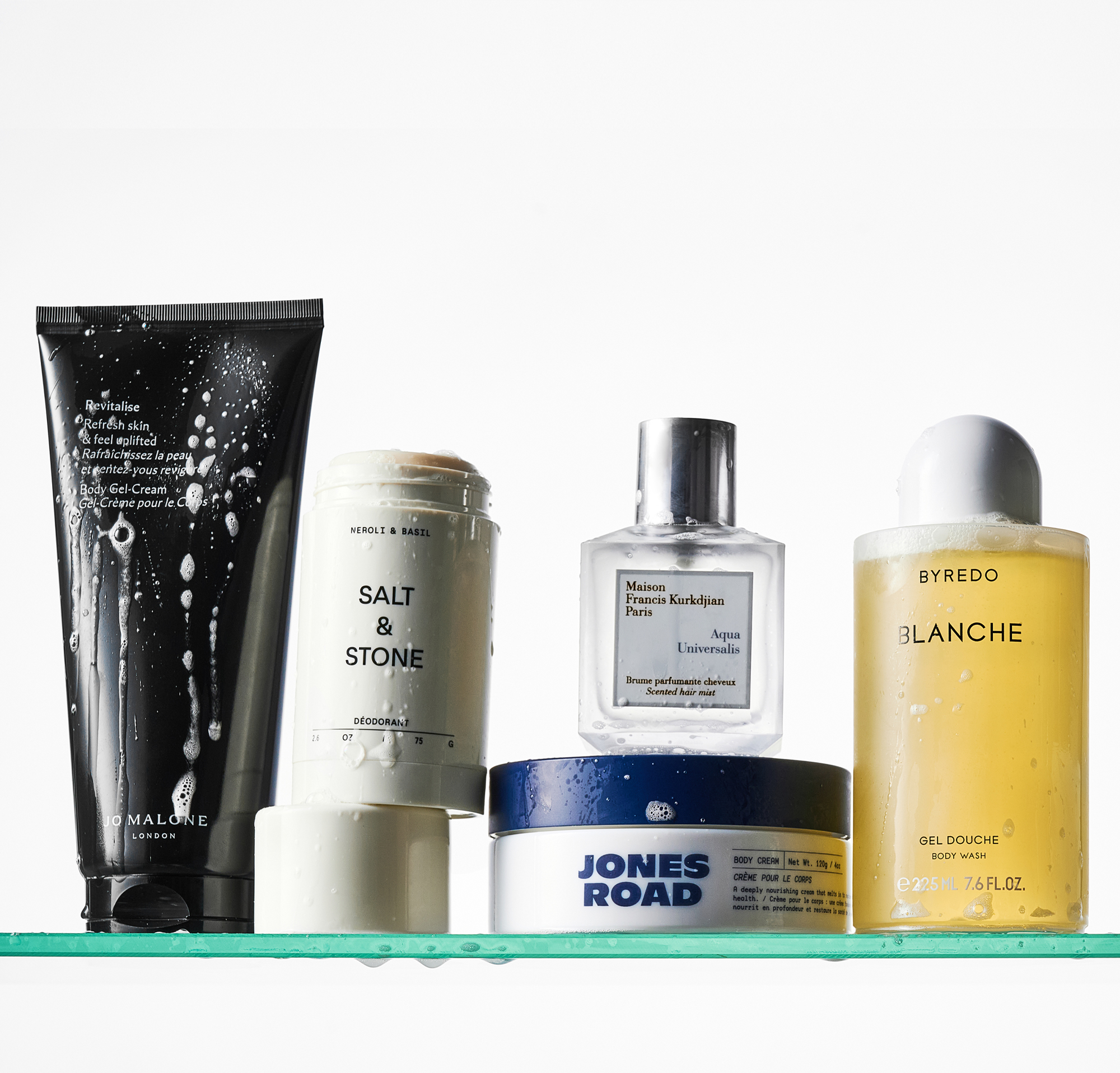 A Jo Malone body lotion, Salt & Stone deodorant, Jones Road body cream, MFK hair mist and Byredo body wash on a glass shower shelf
