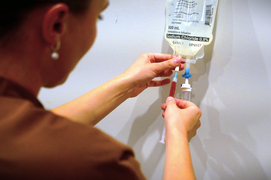A woman, who is turned away from the camera, injects a solution into an IV drip bag.