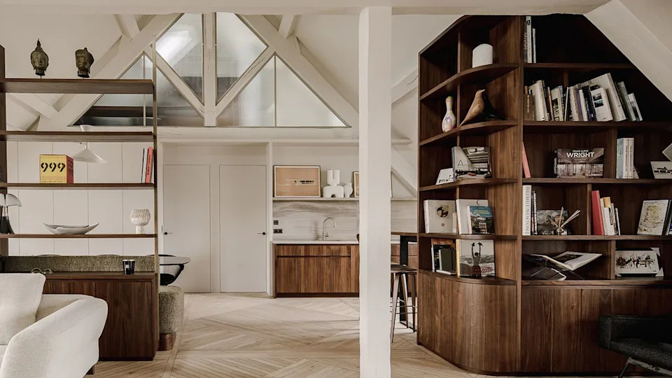  A refined attic apartment interior characterized by a warm, modernist aesthetic. The space is organized as an open-plan living area under a pitched roof, with exposed white-painted beams and triangular dormer windows that bring in soft natural light. 